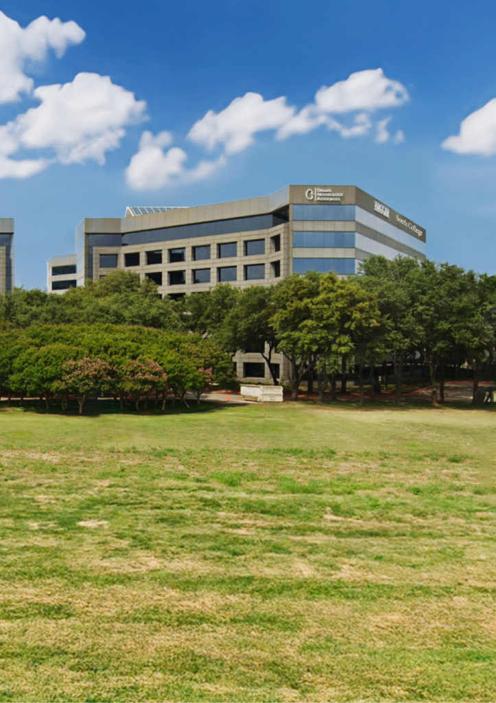 A modern office building with glass windows and "South College" signage, surrounded by trees and a grassy field under a blue sky with scattered clouds.