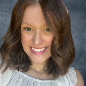 A woman with shoulder-length brown hair and a bright smile poses for a photo against a dark, textured background. She is wearing a white, off-the-shoulder top.