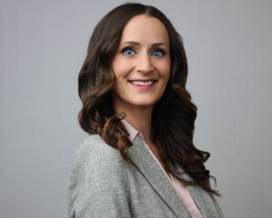 A woman with long, wavy brown hair smiles at the camera. She is wearing a gray blazer over a light pink blouse and stands against a plain gray background.