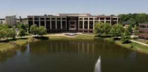 A large, multi-story office building with a modern design stands by a small lake. Trees surround the building, and a fountain is visible in the lake. The sky is clear and blue, indicating a sunny day.