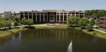 A large, multi-story office building with a modern design stands by a small lake. Trees surround the building, and a fountain is visible in the lake. The sky is clear and blue, indicating a sunny day.