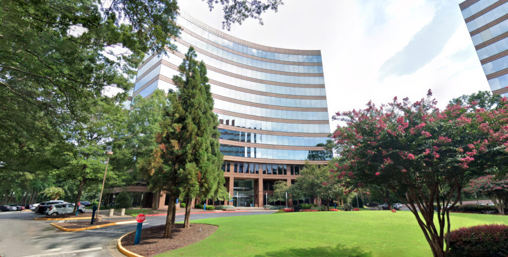 A modern, multi-story office building with reflective glass windows surrounded by well-maintained landscaping, including green grass, trees, and pink flowering shrubs. A paved pathway leads to the entrance.