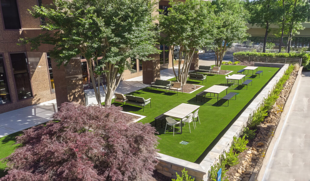 Outdoor seating area with tables and chairs on a grass-covered patio surrounded by trees and shrubs. The area is adjacent to a building. Paths lead through well-maintained landscaping under a clear sky.