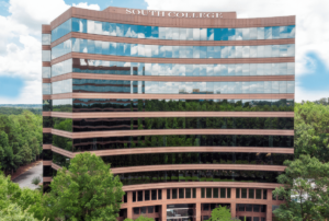 A multi-story building with "South College" on the roof reflects clouds and sky in its glass windows. It's surrounded by lush green trees and set against a bright blue sky with scattered clouds.