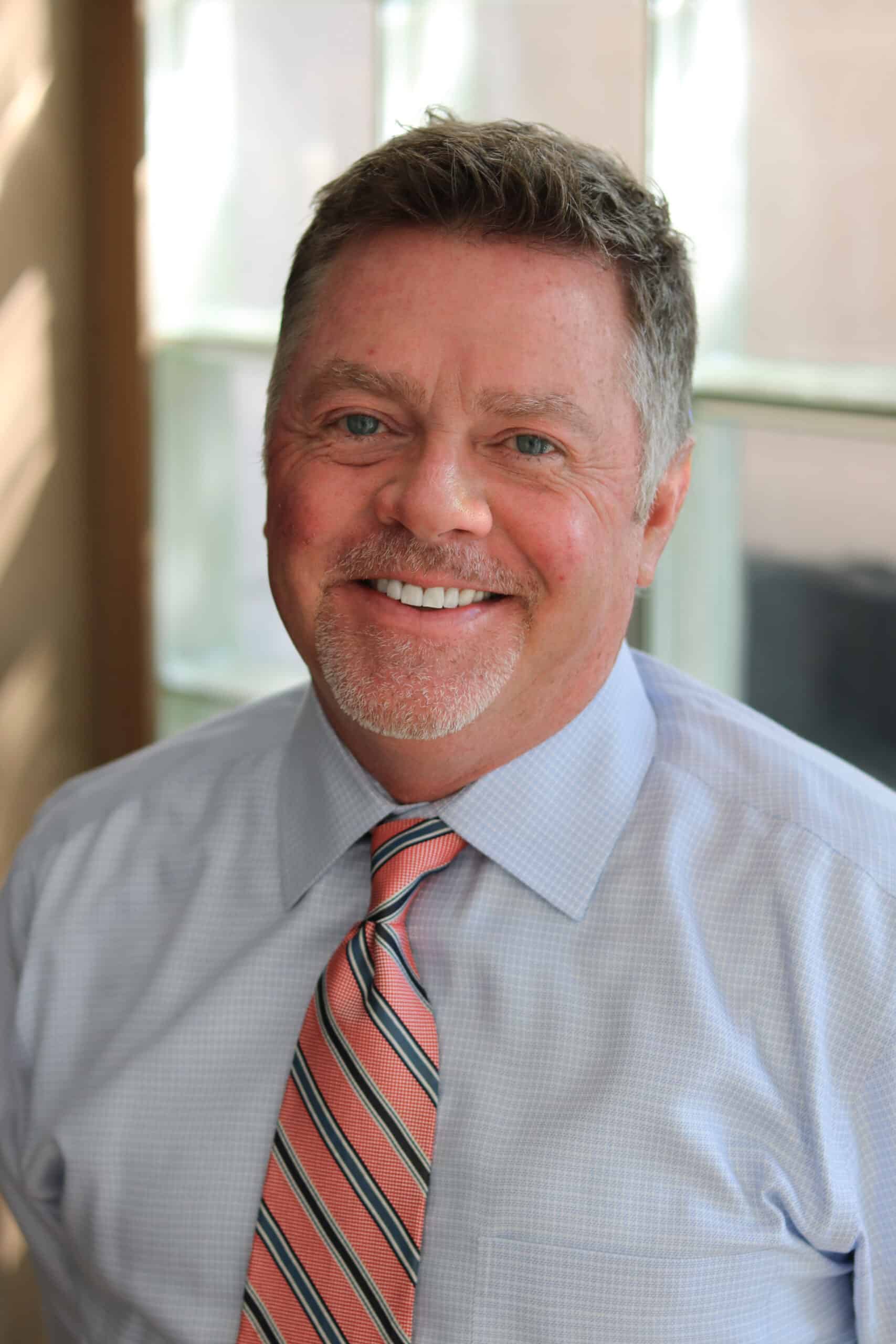 A man with short hair and a beard is smiling. He is wearing a light blue dress shirt and a striped tie, standing in front of a window with soft natural light illuminating the scene.