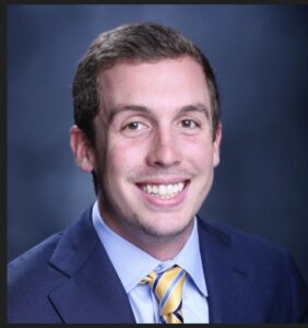 A man in a blue suit and yellow striped tie smiles against a dark blue background. He has short brown hair and light facial hair.