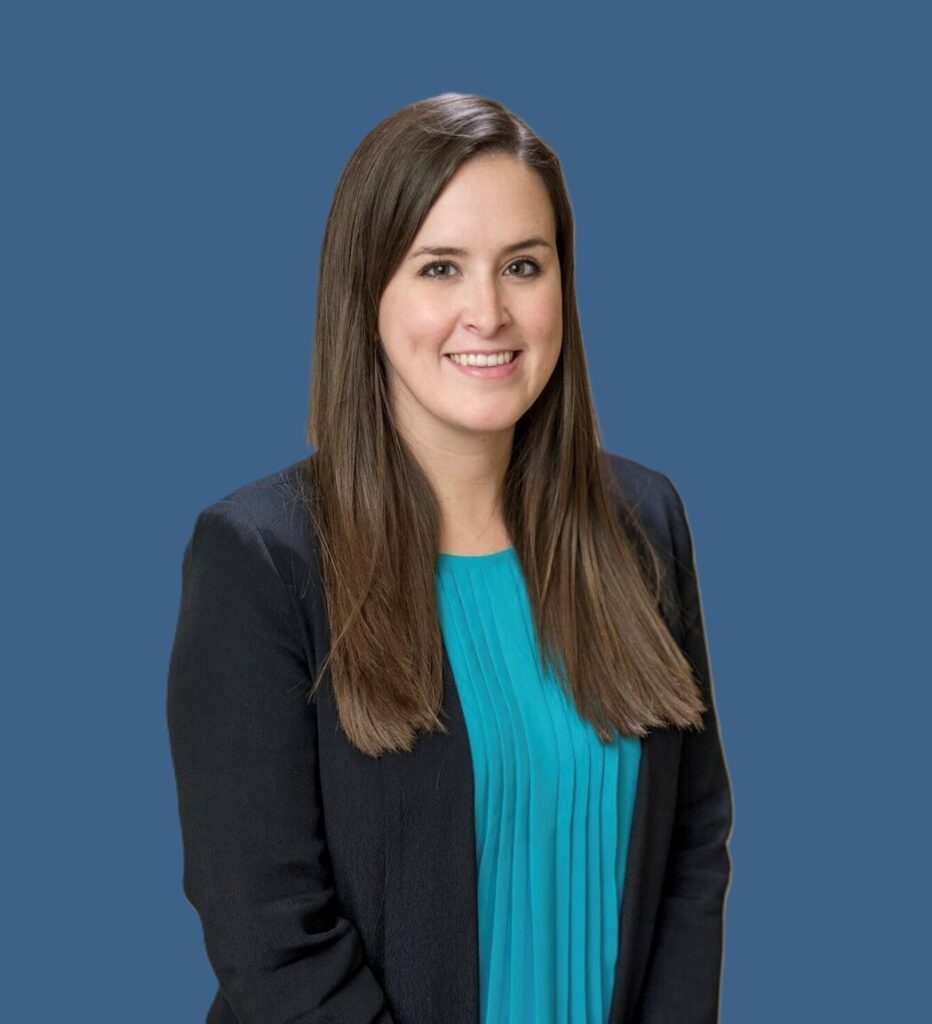 A woman with long brown hair wearing a teal blouse and a black blazer stands against a plain blue background, smiling at the camera.