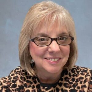 A person with short, light hair and glasses smiles at the camera. They are wearing a leopard print top and are set against a plain background.