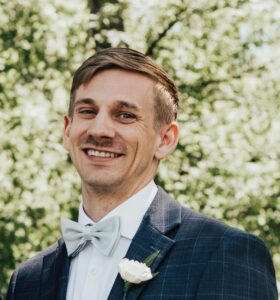 A man in a blue checkered suit and gray bow tie smiles warmly.