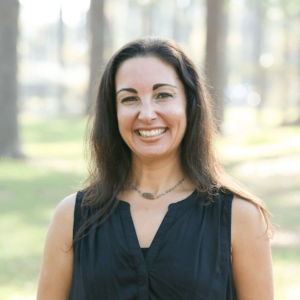 A woman with long brown hair and a black top smiles while standing outdoors in a sunlit park with trees.