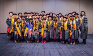 A large group of graduates wearing black gowns and caps, with various colored stoles and tassels, stand together smiling in a conference room. The background is a plain beige wall.