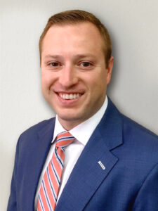 A person wearing a blue suit, white shirt, and a striped red and white tie is smiling at the camera. They have short, light brown hair and are standing against a plain light-colored background.