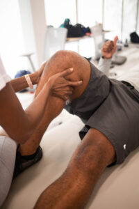 A person is receiving physical therapy in a bright classroom setting. The focus is on their knee, with a physical therapy student's hands applying pressure.
