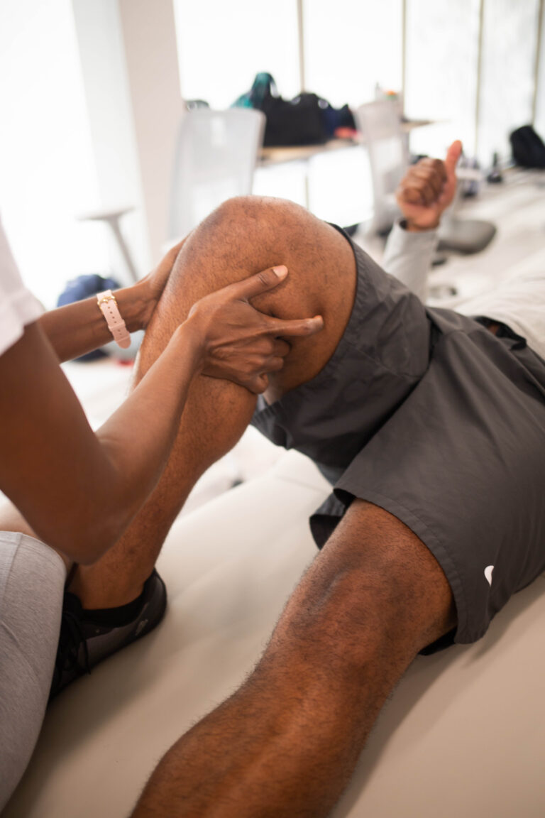 A person is receiving physical therapy in a bright classroom setting. The focus is on their knee, with a physical therapy student's hands applying pressure.