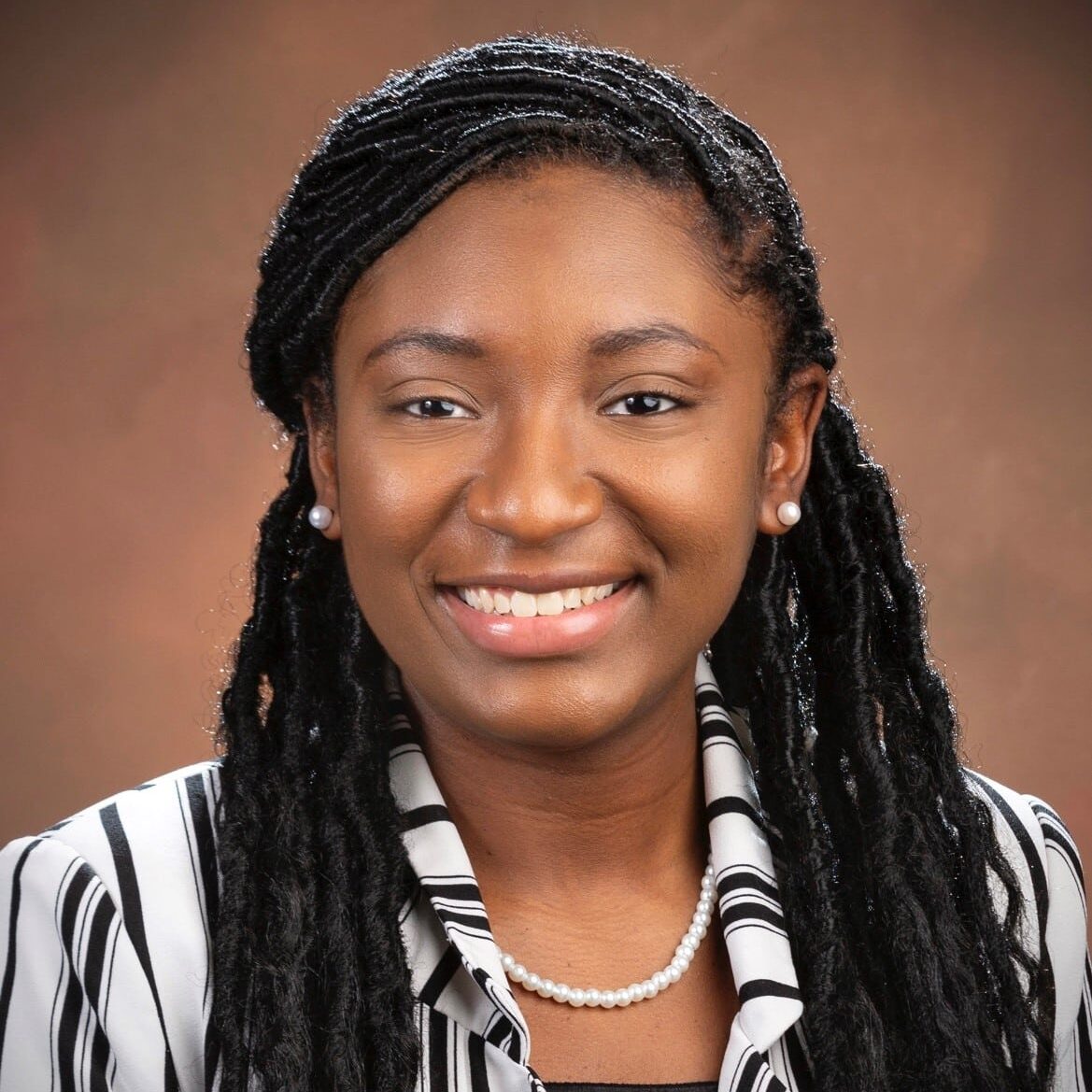 Smiling person with long, braided hair and pearl earrings. Wears a striped blouse and a pearl necklace.