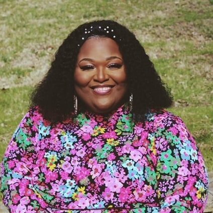 Person sitting outdoors on a stone bench, smiling. They have long, curly hair and are wearing a vibrant floral dress.