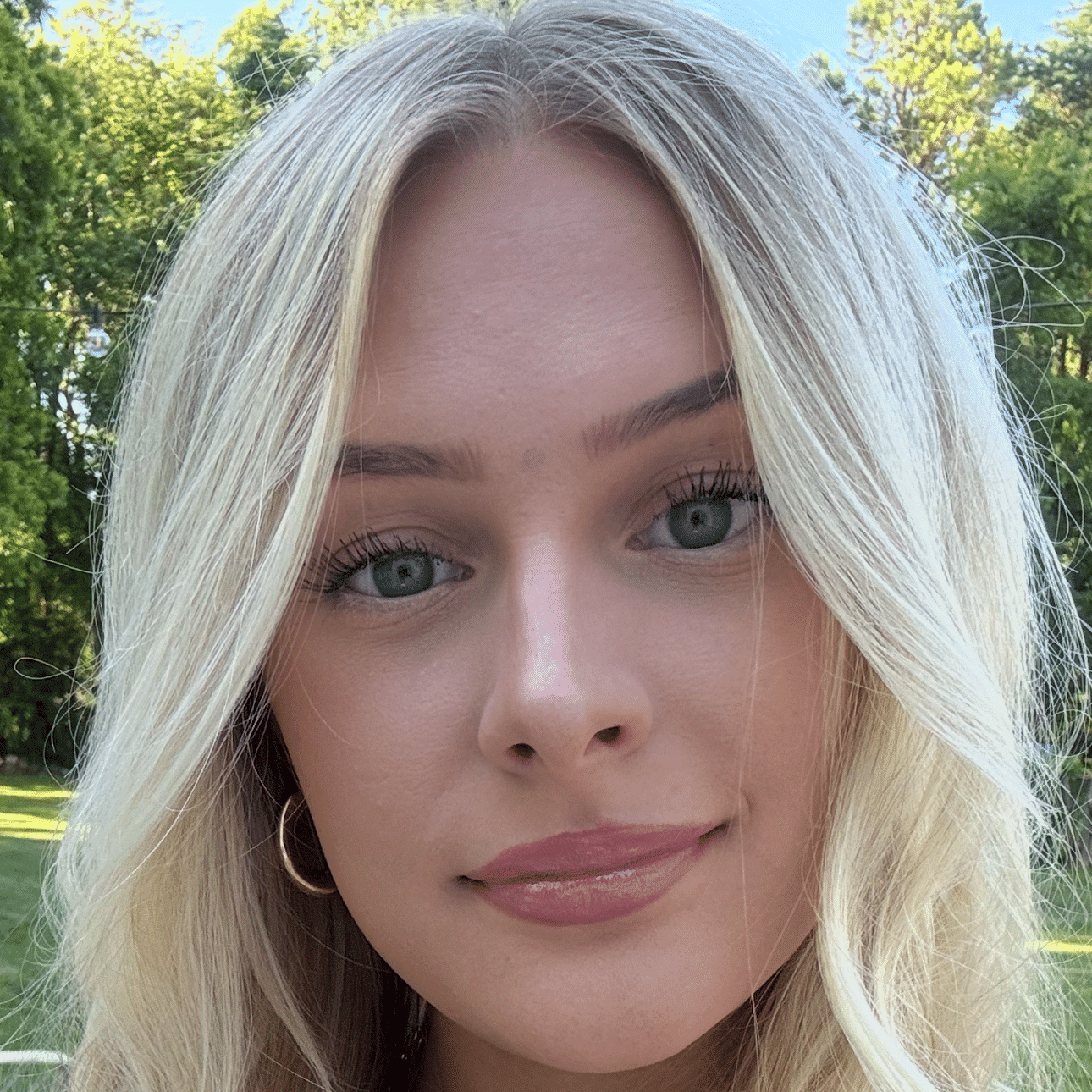 A woman with long, wavy blonde hair is smiling softly at the camera. She is wearing a white top and a necklace.