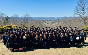 A large group of graduates in black caps and gowns stands outdoors on a grassy area. They smile for the camera with a scenic background of mountains and a clear blue sky. Some hold diplomas, and leafless trees are visible around them.