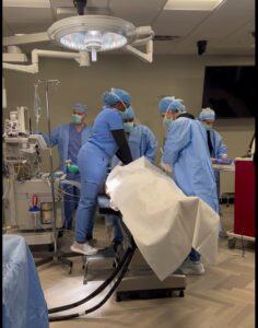 Five medical professionals in blue surgical scrubs and masks are preparing a patient on an operating table. They are gathered under surgical lights in a hospital operating room. Equipment surrounds them, and a large monitor is visible on the wall.