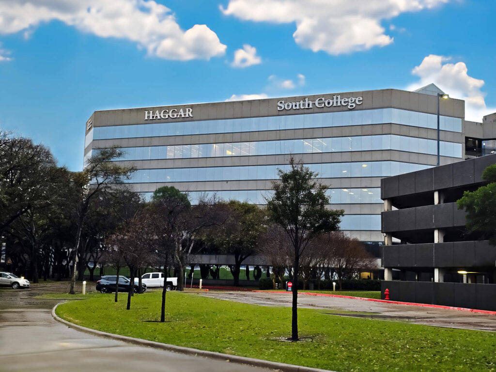 A modern office building with "Haggar" and "South College" on its facade stands against a blue sky with clouds. It's surrounded by trees and a parking area with a white vehicle. Wet pavement hints at recent rain.