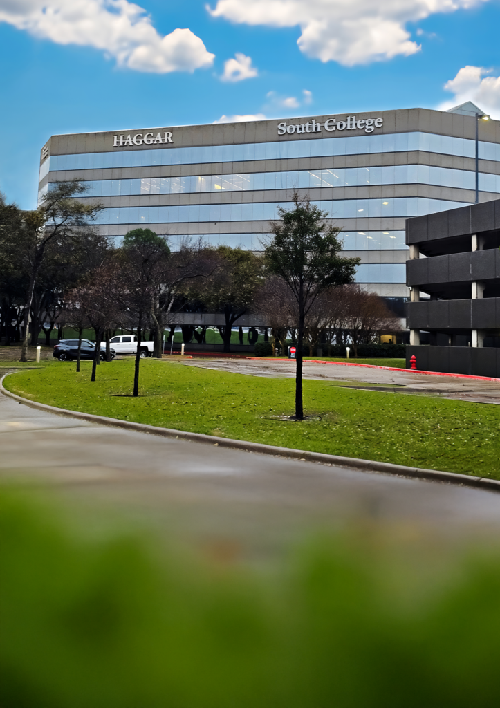 A large building labeled "Haggar South College" with glass windows and surrounded by trees. The sky is blue with clouds, and a grassy lawn is in the foreground with a sidewalk and a few parked cars nearby.