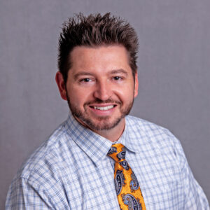 A man with short, spiky hair and a trimmed beard smiles at the camera. He is wearing a light blue checkered dress shirt and an orange tie with blue paisley patterns. The background is plain and gray.