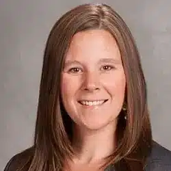 Breanna (Bre) Reynolds A woman with long, straight brown hair smiles at the camera. She is wearing a gray blazer over a pink top, and the background is a plain gray backdrop.