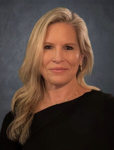 A person with long, wavy blonde hair wearing a black top and a delicate necklace poses in front of a textured dark blue background. They are smiling softly.