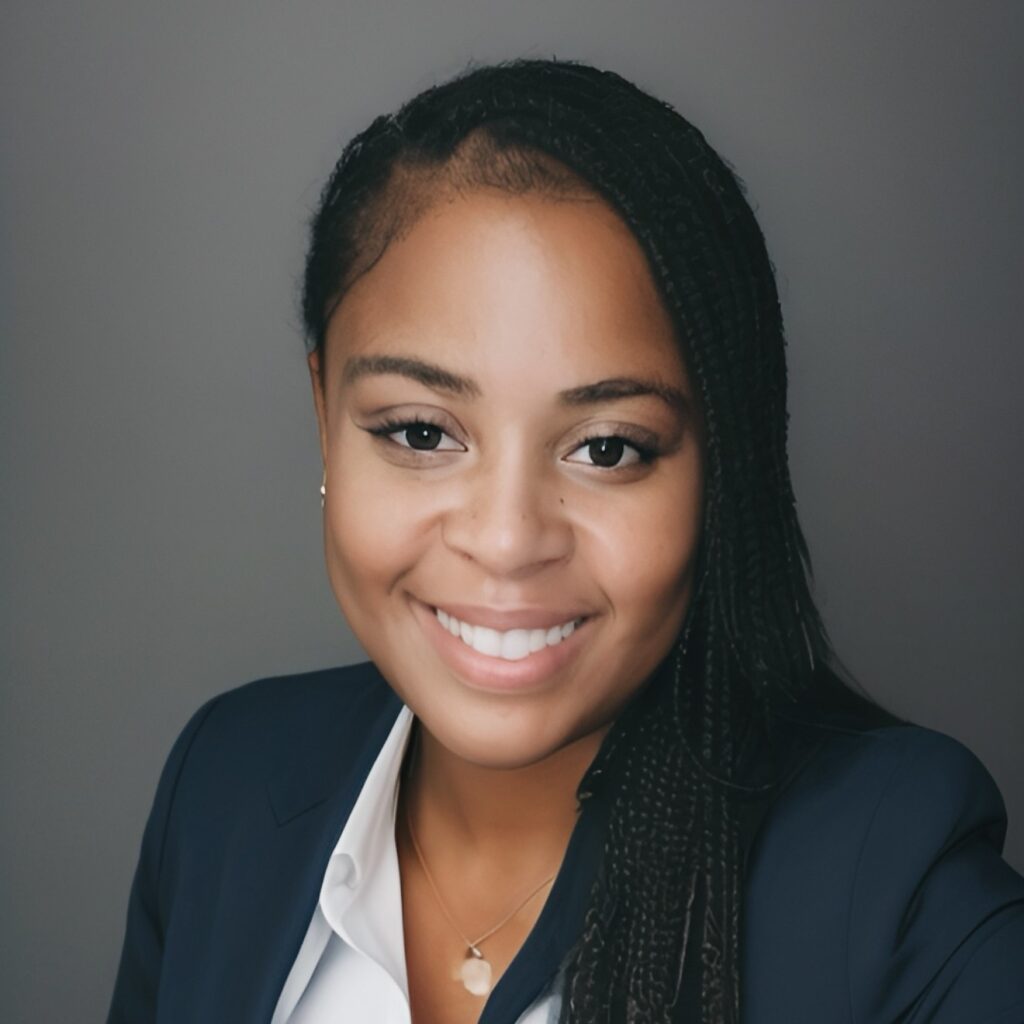A person with long braided hair is smiling, wearing a dark suit jacket over a white shirt. The background is a plain gray.