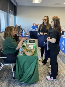 Three women in black scrubs talk to a seated woman at a table covered with a green cloth at a career fair or informational event; brochures, a basket, and water bottles are on the table. Other booths and people are in the background.