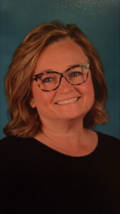 A woman with shoulder-length light brown hair and glasses smiles at the camera. She is wearing a black top and is posed in front of a blue background.