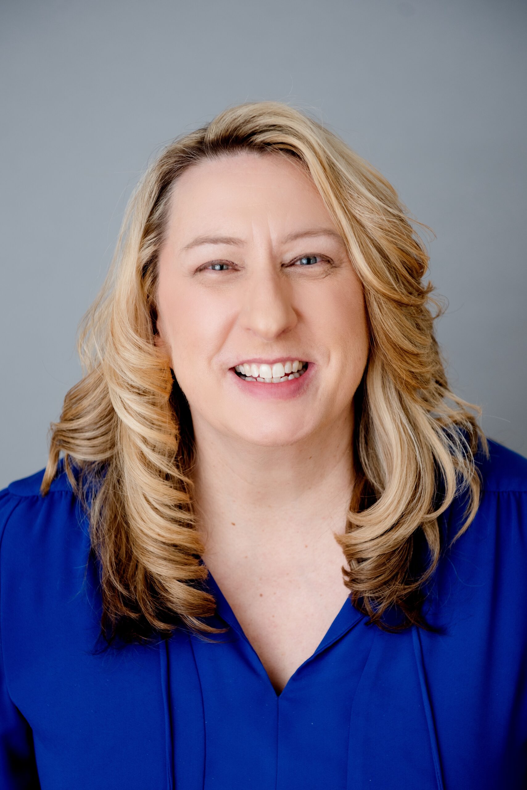 A person with long, wavy blonde hair is smiling at the camera. They are wearing a blue button-up shirt against a plain gray background.