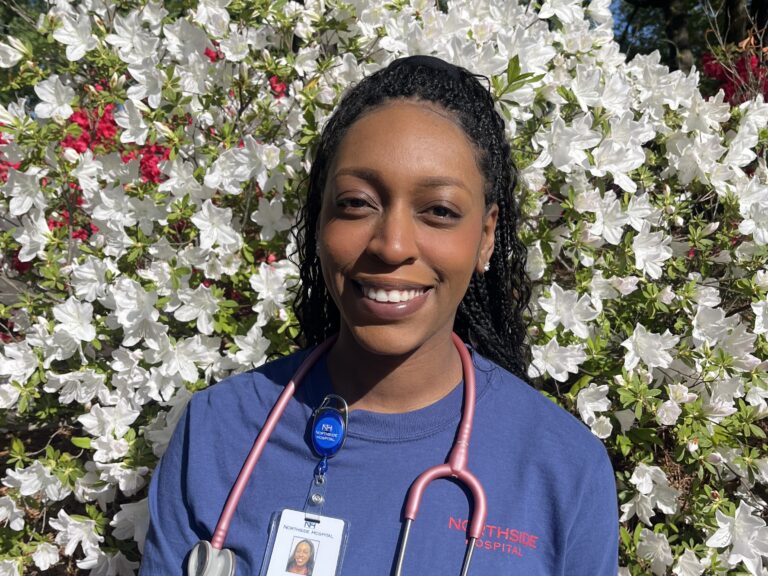 A woman wearing a blue "Northside Hospital" shirt, a stethoscope, and an ID badge smiles in front of white and pink flowering bushes in bright sunlight.