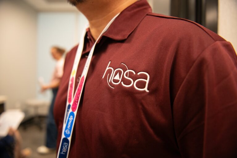 A person wearing a maroon collared shirt with the "hosa" logo and a white lanyard is in focus. The background is blurred, showing another person and a desk.