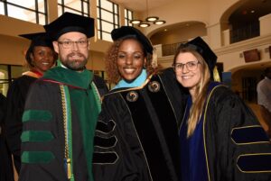Three people in academic regalia smile and pose together indoors at a graduation ceremony, with another person in the background and large windows letting in natural light.
