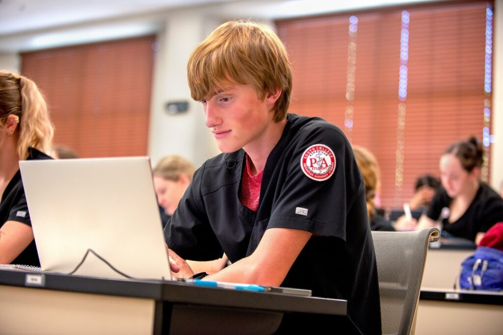 A young student wearing black scrubs with a "Physician Assistant" patch works on a laptop in a classroom, surrounded by other students also engaged in their devices.