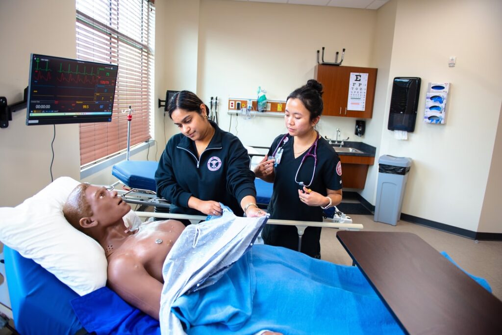 Two nursing students practice procedures on a medical manikin in a hospital simulation room, with medical equipment and monitors visible in the background.