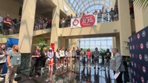 A group of people stand and listen to a speaker in a large, sunlit atrium with a glass ceiling. A red banner celebrating a 10th anniversary hangs above, and people watch from the upper level balcony.