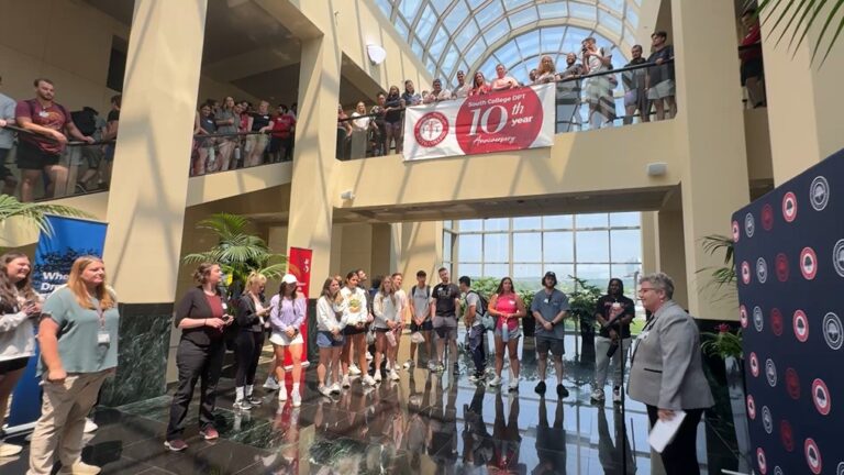 A group of people stand and listen to a speaker in a large, sunlit atrium with a glass ceiling. A red banner celebrating a 10th anniversary hangs above, and people watch from the upper level balcony.