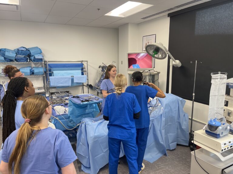 A group of medical professionals in blue scrubs gathers around an operating table, watching a demonstration with surgical tools and a monitor in a well-equipped, bright clinical room.