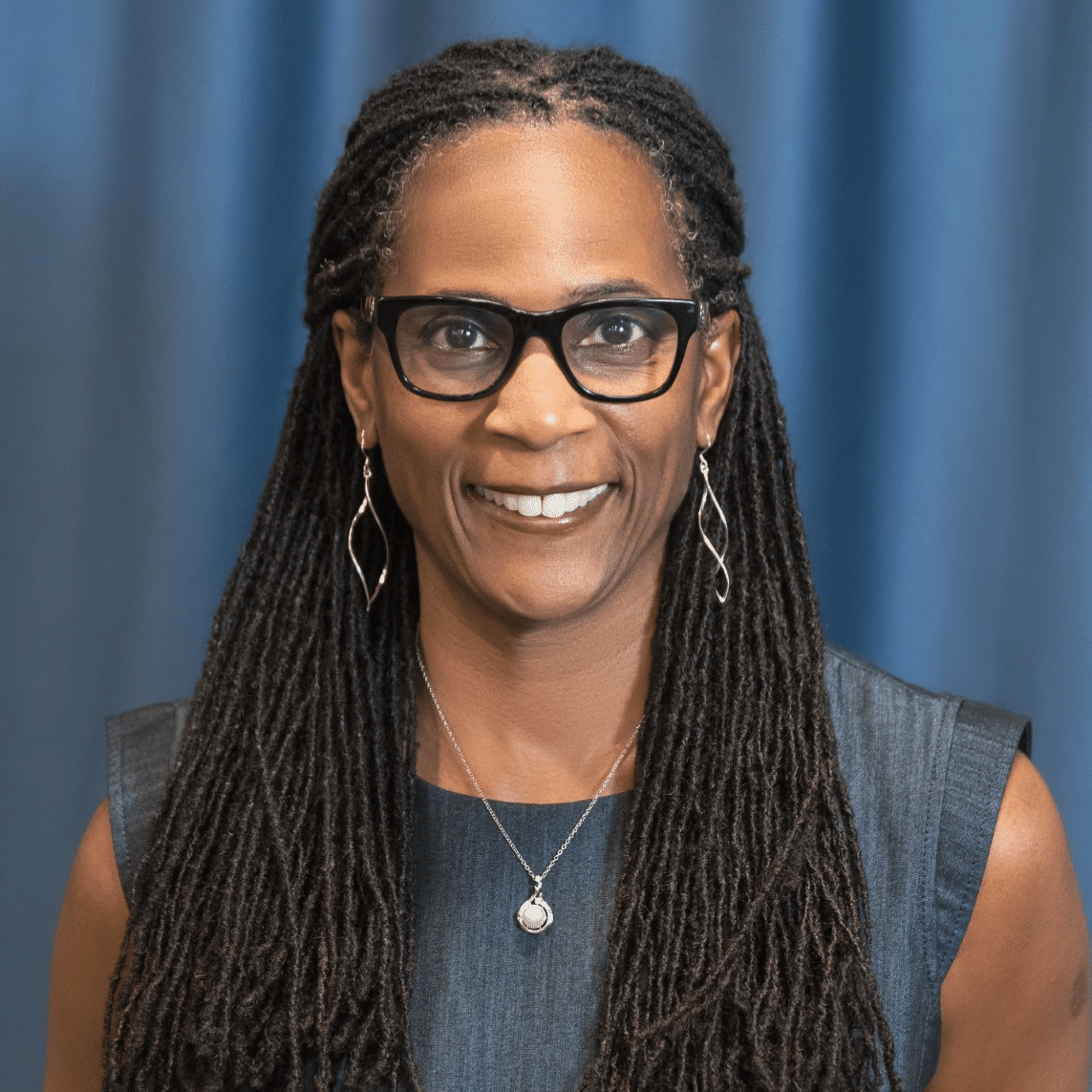 A woman with long, dark, braided hair wears glasses, dangling earrings, and a sleeveless dark dress. She is smiling and standing in front of a blue background.