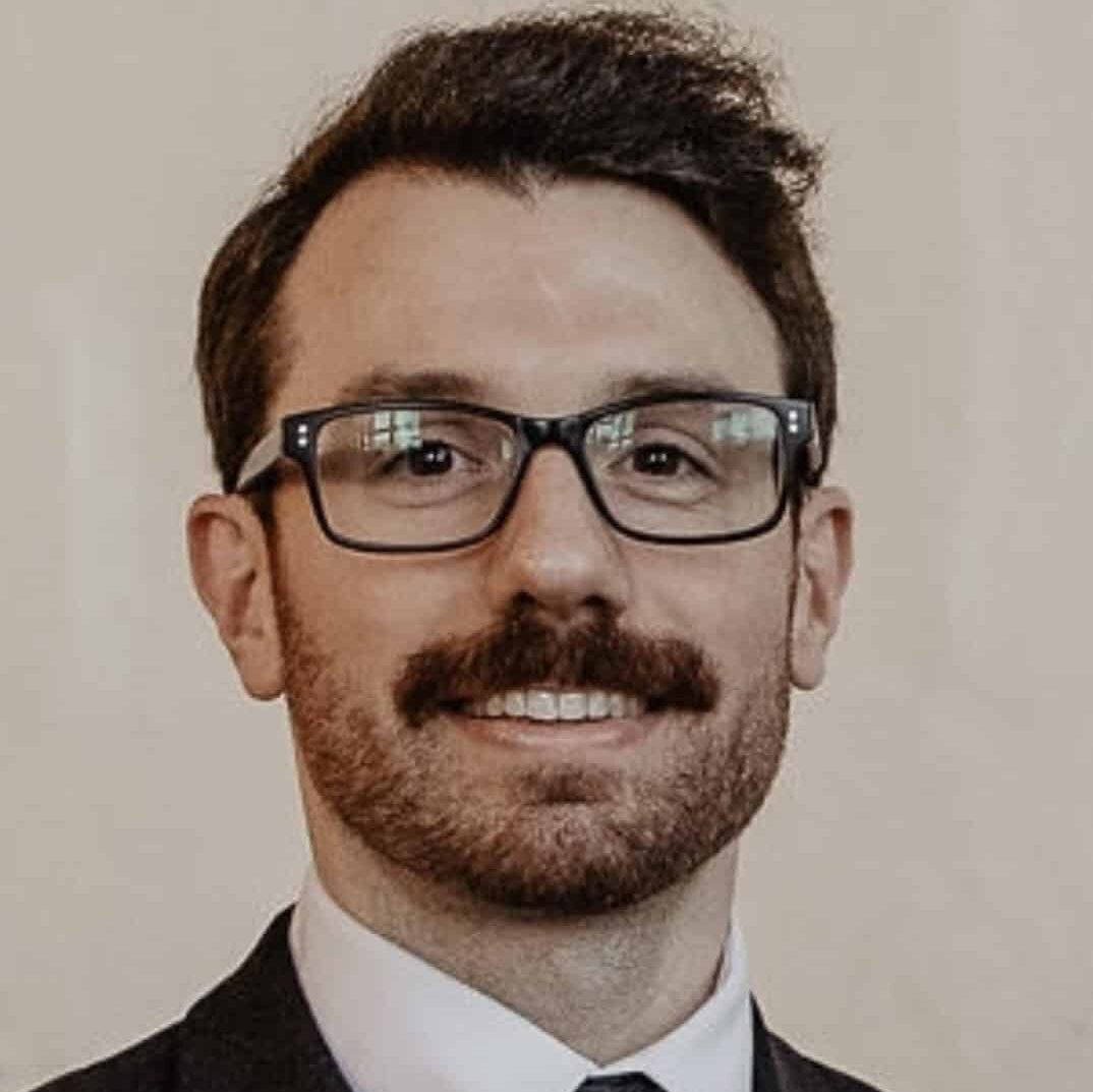 A man with short dark hair, glasses, and a thick mustache wearing a dark suit, white shirt, and patterned tie, smiling in front of a plain light background.