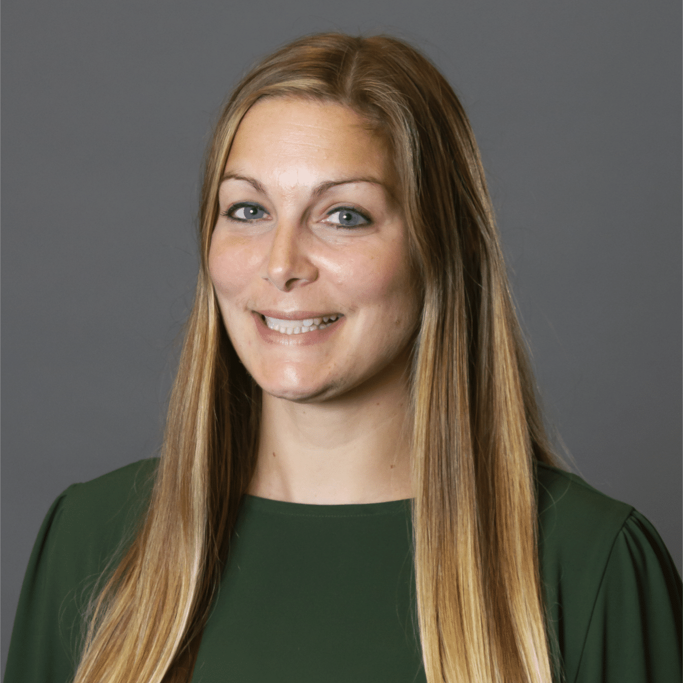A woman with long, straight blonde hair and blue eyes is smiling. She is wearing a dark green top and is posed against a plain gray background.
