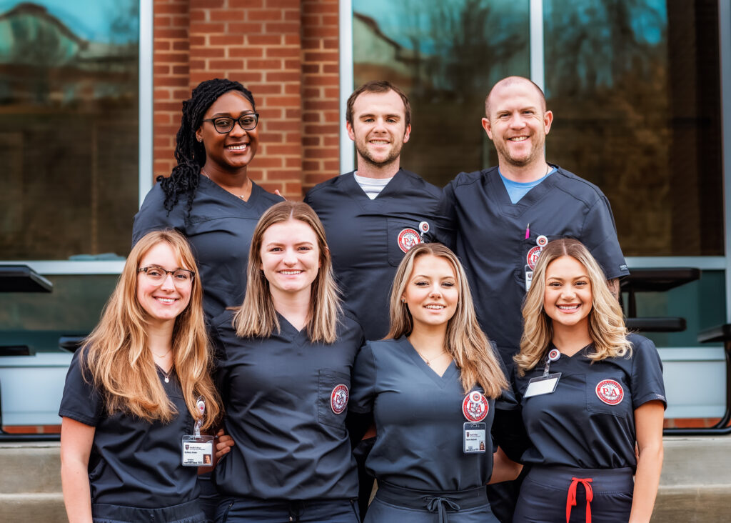 Seven smiling PA students in navy scrubs pose together outdoors in front of a brick building, some wearing name badges and university patches on their uniforms.