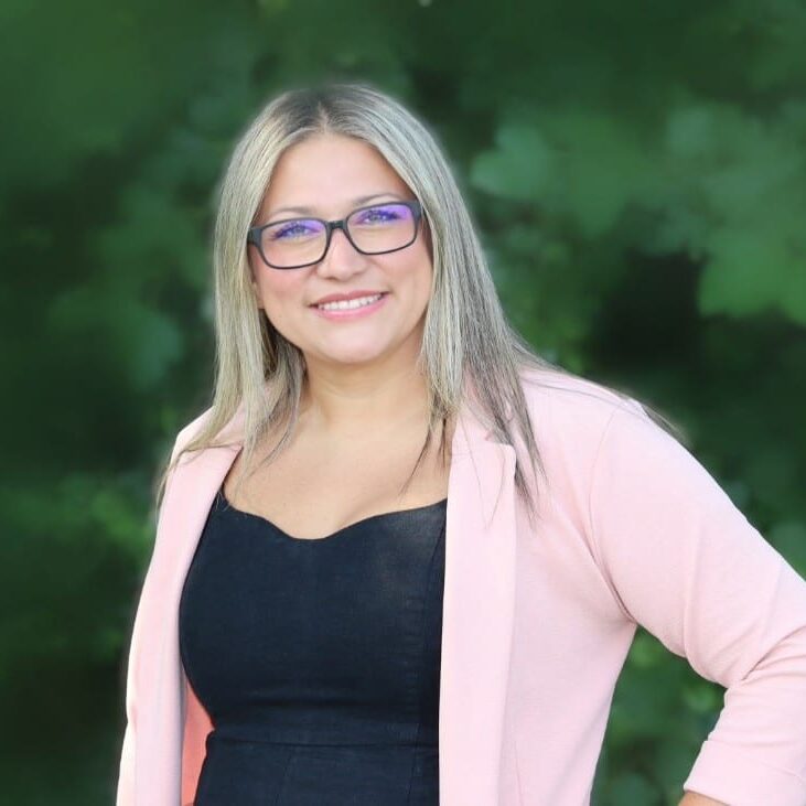 A woman with long, straight blonde hair wearing glasses, a black top, and a light pink blazer stands outdoors, smiling in front of a blurred green background.