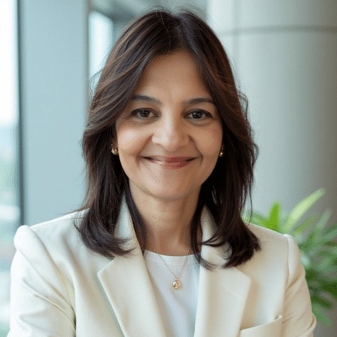 A woman with shoulder-length dark hair, wearing a white blazer and a pendant necklace, smiles softly while standing indoors near a window with natural light and a green plant in the background.