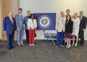 A group of nine professionally dressed people stand smiling beside a sign with the Texas Board of Nursing seal in a well-lit room with a carpeted floor.
