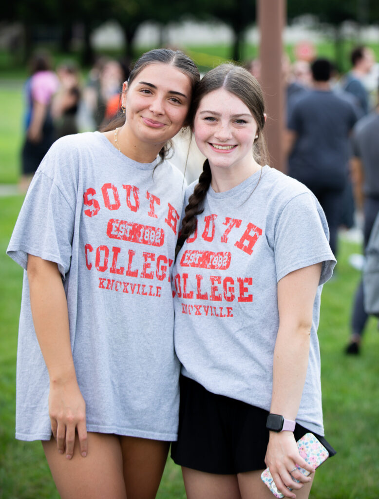 Two young women smile and pose together outside, both wearing gray "South College Knoxville" t-shirts. They stand on green grass with other people blurred in the background.