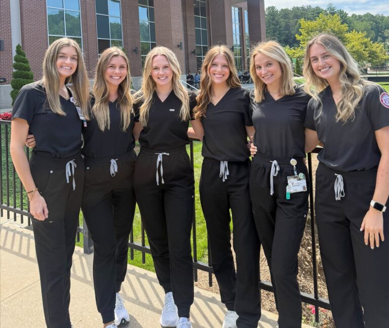 Six women in matching black scrubs stand smiling together outside a building labeled “South College” on a sunny day, with greenery and blue sky in the background.