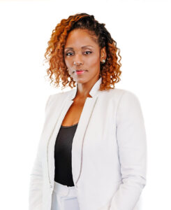 A woman with curly, shoulder-length hair wears a white blazer over a black top. She poses confidently against a plain white background, looking directly at the camera with a slight smile.